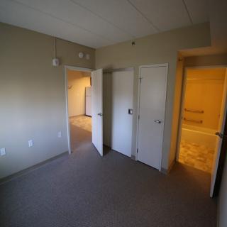 An empty room with carpeted floors, multiple doors, and a view into an adjoining hallway and bathroom.