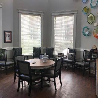 A cozy room with a round table surrounded by chairs, illuminated by natural light from large windows and adorned with colorful wall art.