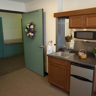 A small kitchenette with a sink, microwave, and refrigerator, adjacent to a doorway leading into a carpeted room.