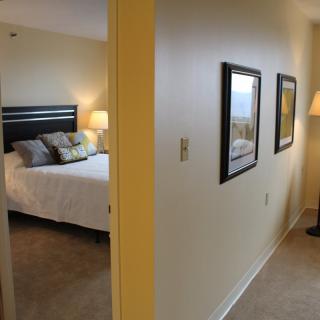 A cozy bedroom is visible from a hallway decorated with framed pictures and a floor lamp.