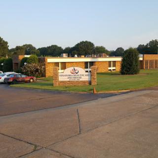 A view of a building with a sign in front, surrounded by greenery and parked cars.