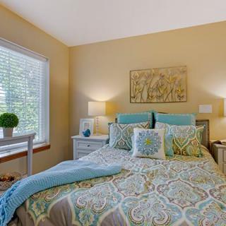 A cozy bedroom with light blue and beige decor, featuring a patterned bedspread, multiple pillows, and natural light streaming through the window.