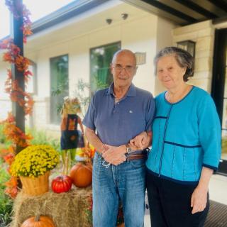 An elderly couple stands together in front of a house decorated for autumn with pumpkins and hay bales.