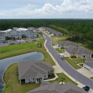 Aerial view of a suburban neighborhood with houses, a road, and a pond surrounded by trees.