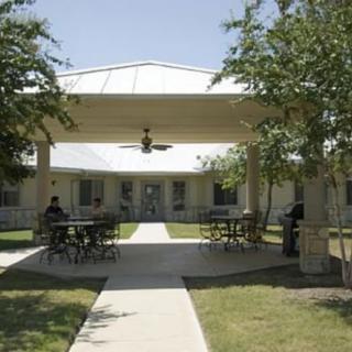 An outdoor seating area with tables and chairs under a covered structure, surrounded by trees and a pathway.