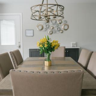 A modern dining room featuring a wooden table with beige upholstered chairs, a stylish chandelier overhead, and a vibrant bouquet of yellow flowers as the centerpiece.