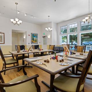 A well-lit dining room with neatly arranged tables and chairs, featuring large windows and elegant chandeliers.
