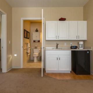 A small kitchenette with white cabinets, a mini fridge, and a microwave next to a bathroom entrance.