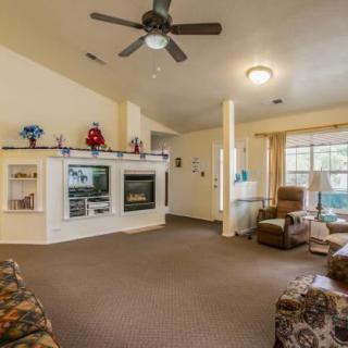 A cozy living room featuring a fireplace, ceiling fan, armchairs, and large windows.