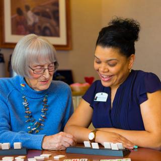 A caregiver assisting an elderly woman with a board game, both smiling.