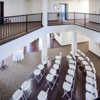 A modern indoor event space with rows of chairs arranged for a conference or meeting, featuring a balcony overlooking the area.