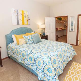 A cozy bedroom with a bed featuring blue and yellow bedding, complemented by a small open closet and decorative wall art.