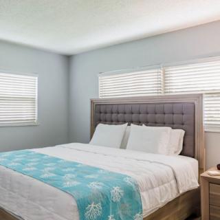 A cozy bedroom featuring a large bed with white and turquoise bedding, illuminated by natural light from two windows with blinds.
