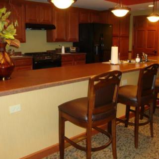 A cozy kitchen with wooden cabinets and a long countertop, featuring bar stools and decorative lighting.