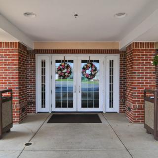 The image shows the entrance of a brick building with double glass doors adorned with decorative wreaths. Trash cans are placed on either side.