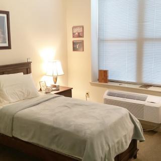 A cozy bedroom with a single bed, nightstand, and lamp next to a large window.