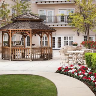 A scenic outdoor garden featuring a wooden gazebo surrounded by colorful flowers and a well-maintained lawn.