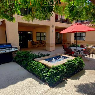 A peaceful courtyard featuring a small fountain, outdoor seating with a red umbrella, and a grill area surrounded by lush greenery.
