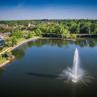 Aerial view of a park featuring a large lake with a prominent fountain at its center, surrounded by lush greenery and city buildings in the background.