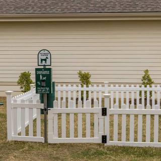 A white picket fence encloses a small yard with a dog waste disposal sign, adjacent to a house.