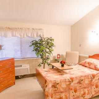 A cozy bedroom featuring a bed with floral bedding, a small nightstand, and a potted plant by the window.