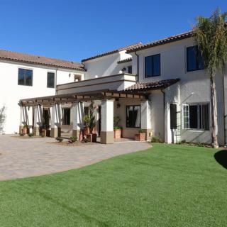 A large, two-story residential house with a well-maintained lawn and palm trees under a clear blue sky.