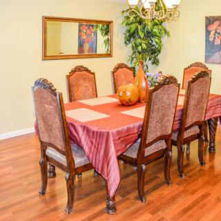 A warmly decorated dining room featuring a wooden dining table with a red tablecloth, surrounded by six ornate chairs. A chandelier hangs overhead and a mirror is mounted on the wall.