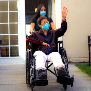 A caregiver pushes an elderly person in a wheelchair outdoors, both wearing masks.