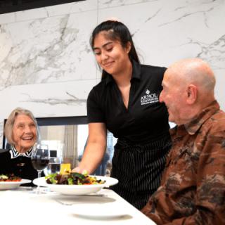 A waitress serves a meal to two seniors in a restaurant setting.