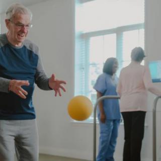 An elderly man engages in physical therapy with a ball under the guidance of healthcare professionals.