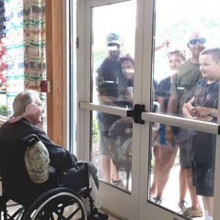A group of people joyfully interacting with an elderly man in a wheelchair through a glass door.