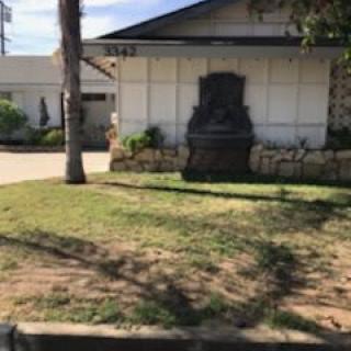 A view of a house with a grassy lawn, a tree, and a statue on a sunny day.