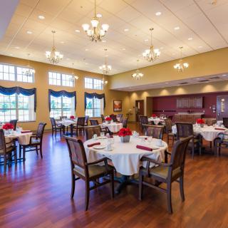 A well-lit dining room with elegantly set tables and chairs, featuring large windows and chandeliers.