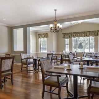 A well-lit dining area with tables and chairs, featuring a chandelier and large windows.