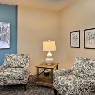 A cozy living room corner with patterned armchairs, a lamp on a wooden side table, and framed art on the walls.