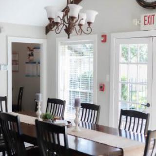 A bright dining room with a long table, chairs, and a chandelier, illuminated by natural light from the windows.