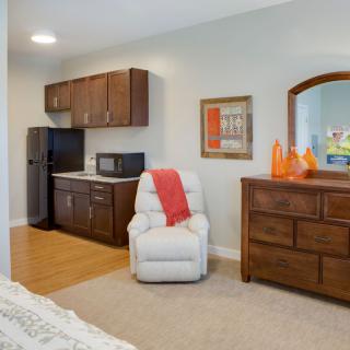 A cozy bedroom featuring a small kitchenette, a reclining chair, and a wooden dresser.