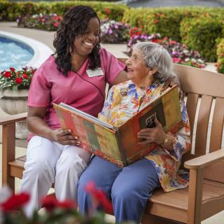 A caregiver and an elderly woman share a book together while sitting on a bench in the garden.