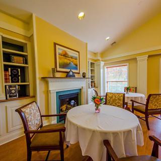 A cozy dining area with round tables, chairs, a fireplace, and a bookshelf in a brightly lit room.