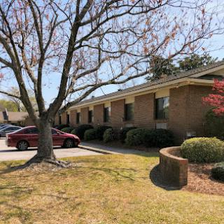 A beautiful day with clear skies, showcasing a residential building surrounded by greenery and a parking area.