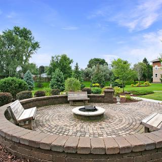A beautifully landscaped outdoor space featuring a circular brick patio with a central fire pit and surrounding wooden benches.