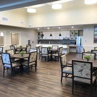 A modern dining area with a kitchen, featuring tables and chairs arranged on polished wooden flooring.
