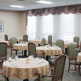 An empty dining room with round tables and chairs arranged neatly, featuring large windows covered with curtains.