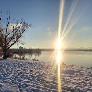 A tranquil winter evening by the lake, with snow blanketing the ground and the sun setting beautifully on the horizon.