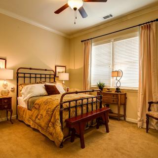 A cozy bedroom with warm lighting, featuring a bed with decorative pillows, bedside tables with lamps, and large windows with curtains.