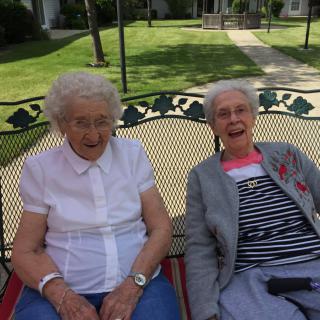 Two elderly women sitting and smiling on a bench in the park.