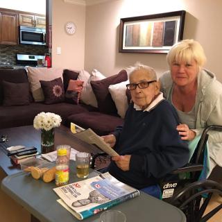 An elderly person sitting in a wheelchair reading a magazine, accompanied by another person in a cozy living room.