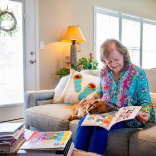 A woman sits on a couch in her living room, reading a book while her dog rests beside her.
