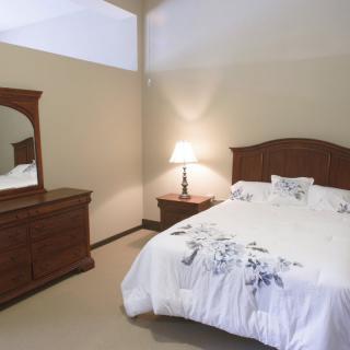 A cozy bedroom with a wooden bed, dresser, and nightstand, lit by a table lamp.
