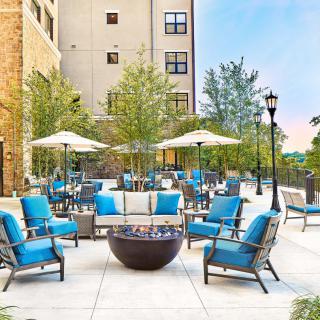 A modern outdoor patio featuring blue cushioned chairs, umbrellas, and a central fire pit in an urban environment.